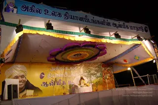 Night photo of an outdoor stage under a white canopy with yellow and green fringe and a large circular floral-style decoration in purple, green and yellow. A seated figure in a white cushioned chair on a white draped platform is wearing yellow and white robes; the figure is small in the distance and its face is not clearly visible. A large yellow banner with a portrait labelled with a Tamil name in the lower left shows a smiling man in a white turban and gold necklace, and the banner carries Tamil text announcing an all-world Dhyanapeetam spiritual conference. Above the stage is a blue banner with Tamil text and a circular logo. Stage lights hang from the canopy frame and a speaker stands at the left edge. The seated figure is a speaker at the event; whether the figure is the person pictured on the banner is uncertain.