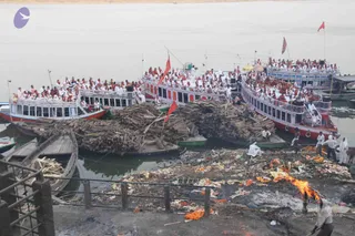 Photo from a riverside ghat on a wide brown river: several white and red passenger boats are packed with people in white and saffron clothing, many standing and seated along their rails. In the foreground stacks of cut firewood lie on the bank beside a burning pyre, with orange cloth and flower offerings scattered on the ground, and a man in white working near the flames. Red flags fly from the boats and a yellow umbrella shades a deck. Several figures in white stand on the steps and shore. The scene suggests a cremation ghat with boats carrying devotees.