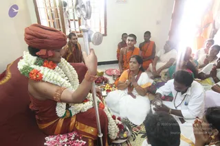 A padapuja ritual in a small room with cream walls and a window. A seated figure in a rust-red turban and red-orange robe, wearing white and orange flower garlands and orange beaded bangles, holds a long silver staff upright and raises a hand. Before him a woman in white sari with an orange border sits with palms joined in prayer, flanked by offerings of fruit, a coconut, red flower petals and a brass plate. Around them saffron-robed young boys with shaved heads and rudraksha malas sit on the floor, and white-clad devotees, including older women and a man in white shirt holding a small brass vessel, watch or join in.