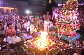 Night outdoor fire ritual with a large square homa pit of stacked brick-like altar, lit by a bright flame, decorated with red flower petals and white rangoli-style borders. A priest in white dhoti with a green headcloth and green shawl stands tending the fire with a long ladle, while a shirtless man in white dhoti beside him holds his hands in anjali. A seated man on the left, bare-chested with a white dhoti and gold necklace, holds a small bell. Behind them, a large ornate chariot or ratha with garlands, carved lions and a golden canopy stands on a tiered base. Many seated devotees in white and red shawls watch from the left. Brass pots, fruit offerings and a plate of flowers sit near the fire.
