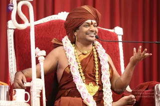 A man seated on an ornate white and silver throne with red upholstery, against a red curtain backdrop. He wears a rust-brown turban, a brown-orange robe draped over one shoulder, long gold beaded malas and a gold pendant, and pink-and-white flower garlands. He smiles and gestures with his right hand while speaking into a microphone on a stand; a white cup sits on a side table.