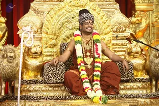 A photograph of a smiling man seated on an ornate golden throne carved with lion heads, with leopard-print cushions and a leopard-print headwrap. He wears a rust-brown dhoti-style garment, a multicoloured garland of white, yellow, green and red flowers, gold necklaces and armlets. A white ceremonial staff with a snake-like carving leans at left, and a microphone stands at right. The background shows gilded carvings and red drapery.