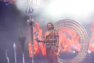 A man stands on a stage in front of a large ornate silver-gold circular frame, with orange flames painted on the backdrop behind him and smoke drifting in the lighting. He wears a leopard-print bodice with rows of brass bells across the chest and waist, a rust-coloured dhoti, a rudraksha bracelet, and a thin neck chain, with long dark hair falling to his shoulders. He holds a silver trident in one hand. To the left are a silver trident on a staff, a drum bound with a leopard-print cloth, a peacock-feather whisk, and a silver staff with a finial, all arranged as stage props.