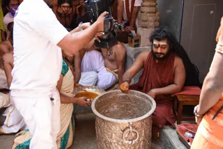 A photograph of a temple ritual in progress. A bearded man with long dark hair, a white forehead marking and a red-orange robe sits on a low stool and pours orange powder into a large ornate brass vessel carved with relief figures. Beside him a woman in a cream sari with teal border holds a brass plate of the same orange powder. A man in a white shirt films with a video camera at left, and several bare-chested men in white dhotis and onlookers sit behind. A stone pillar with carved designs stands at upper centre.