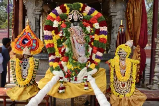 A decorated deity murti stands at the centre of a temple shrine, wrapped in a large garland of red, pink, white and purple flowers with a yellow marigold border, and draped in a golden-yellow cloth. The central figure wears an ornate jewelled breastplate and a decorated crown. On either side, smaller murtis wear yellow marigold garlands and bead necklaces; the left figure is under an orange canopy with a dove emblem. White ceremonial cloth strands are held at the base, and a red-and-gold cloth hangs behind. A devotee in a dark shirt stands at the left edge with hands folded. No living a person is visible; the figures are statues.