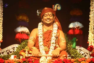 A seated man in saffron-orange robes sits on an ornate silver throne decorated with red and pink roses and green leaves. He wears a saffron turban with a peacock feather ornament and a red tilak on his forehead, a white and pink flower garland, a rudraksha mala, gold necklaces and gold armbands. He holds a gold-and-silver ceremonial sceptre across his lap and smiles with eyes closed. Behind him hang tiered tassel decorations in red, white, purple and yellow, with flower strands on either side and a small white cup at lower left. Swamiji (probable): the saffron robes, peacock feather turban, rudraksha mala and ornate gold jewellery are the qualifying cues for the seated central figure.