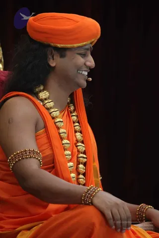 A photograph of a smiling man seated in profile, wearing an orange turban with a gold-trimmed edge, an orange shawl draped over bare shoulders, and a long multi-bead gold-and-black necklace. He wears a small microphone headset and beaded rudraksha-style bracelets on his arms and wrists, and his hands rest in his lap against a dark maroon curtain backdrop. A small red tilak mark is visible on his forehead. The lower right corner carries a copyright notice. Based on his orange turban, saffron-toned draped clothing and layered gold-and-rudraksha adornments, this is a Swamiji (probable) appearance-based label, seated on stage as a speaker.