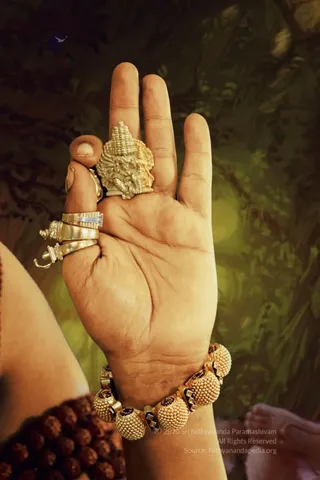 Close-up photograph of a bare hand held palm-up in front of a dark, swirling green-and-brown painted backdrop. The palm holds a small ornate gold deity murti with a tiered crown and multiple arms, resembling a Ganesha-style figure. Several gold rings are worn on the fingers, including a band set with white stones. A heavy gold bracelet of beaded spheres with dark enamel inlays is on the wrist. Dark rudraksha beads and a sleeve of pale fabric are visible at the lower left edge, and a blurred figure appears at the lower right.
