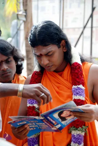 Photograph of a long-haired young man in saffron-orange robes wearing a red and white garland with purple flowers and a red bindi on his forehead, who is looking down at a blue printed brochure showing a smiling woman's portrait while another person's hand reaches into the frame. Behind him a second man in orange with a red forehead mark watches. Outdoor setting with a palm tree and building in the background. Appearance-based candidate: Swamiji (probable), long swept-back hair and saffron robes with a flower garland, present in person.