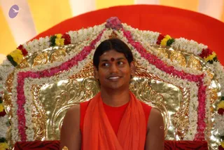A man in saffron-orange robes sits on an ornate gold throne decorated with garlands of white jasmine, pink and red flowers, and yellow marigolds, with a lotus-shaped finial at the top. He has a red bindi on his forehead and smiles while looking off to one side. Behind the throne are bright yellow and orange striped fabric drapes. The scene resembles a formal satsang or darshan setting.