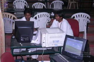 A photograph of two young men in white clothing seated at a table in a hall with rows of white plastic chairs. The man on the left, with short dark hair and a moustache, works at a beige CRT monitor, while the man on the right, wearing glasses and a red-orange lanyard, looks toward the screen beside a white HP laser printer. A second monitor showing a blue-green desktop wallpaper sits in the foreground, with papers and cables on the table.