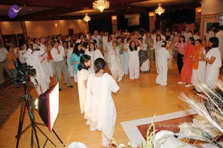 A large hall with a wooden floor and crystal chandeliers shows a crowd of dozens of people, mostly dressed in white, standing and clapping with raised arms in a circle-like group. Many wear white kurtas, pants and saris, with a few in orange, pink and teal. A camera on a tripod and a studio light box stand at left, and a peacock feather arrangement and a small rug with white cloth are at the bottom right. A framed banner hangs on the back wall. Several people in the foreground hold their hands up in a devotional clapping gesture.