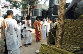 A street-side photograph beside a large gold-coloured wooden chariot (ratham) with carved floral and bird relief panels. A man in a white dhoti and white shirt holds a camera on a shoulder rig, filming. In the centre, a man with long dark hair wears an orange saffron robe and a white-and-red flower garland, standing barefoot before the chariot. Beside him, an older man in white clothes with hands folded in prayer, and two men in white dhotis with orange shawls stand nearby. Black water storage tanks and a tree stand behind them on a dirt road.