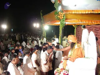Night outdoor event with a crowd of many people, mostly men and women in white shirts, white sarees and white dhotis, some wearing lanyards, gathered before a stage. On the stage, a seated figure in a saffron-orange turban and saffron-orange garment wears a long garland of white and orange flowers and beads, and extends an arm toward a man in a white shirt who is bowing with hands pressed together. Behind the seated figure stands another man in white. The stage has an orange and white canopy with a bunch of green coconuts and banana stalks hanging from the pole, a maroon draped backdrop, and floodlights illuminating the scene.