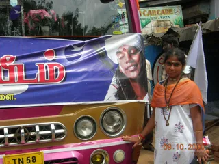 A photograph of a woman standing beside the front of a pink and gold bus parked on a roadside. She wears a white salwar kameez with blue floral print, an orange shawl draped over one shoulder, a long rudraksha-style bead mala and a gold pendant, and bangles on her wrist. A large blue banner on the bus front shows a portrait of a man with long dark hair, a white head covering and a red tilak, with Tamil lettering in red. Behind her a man in a dark shirt stands near a roadside shop with a 'CAR CARE' sign and a white flag. A yellow Tamil Nadu number plate reading TN 58 is visible on the bus.