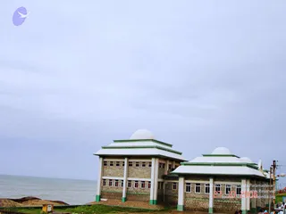 A photograph of a two-storey, brick-and-concrete building with white domed roofs and green trim, standing on a sandy shore beside a grey sea under an overcast sky. Two or three small figures are visible at the lower right near the building's base. A utility pole and a small yellow box sit at the lower left.
