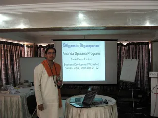 A photograph of a business development workshop held in a hall with heavy floral curtains and ceiling lights. A man in white kurta and white trousers with an orange shawl over his shoulders and a rudraksha mala stands at left holding a handheld microphone. Behind him a projector screen shows slides reading Nithyananda Dhyanapeetam, Ananda Spurana Program, Parle Foods Pvt Ltd, Business Development Workshop, Daman, India, 2006 Dec 21, 22. A laptop sits on a draped round table at right, with a flipchart and a sign reading Cidan... Beau... at the far right edge. Books and papers lie on a side table at left.
