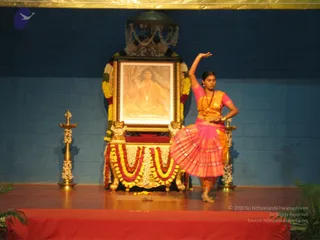 A female dancer in a pink and orange silk costume performs on a stage in front of an ornate golden throne draped with yellow, orange and red marigold garlands and white flower strings. A framed portrait labelled "Paramahamsa Nithyananda" sits on the throne, showing a seated figure in orange robes with a rudraksha mala. A tall gold oil lamp decorated with flower garlands stands on each side, and the blue wall behind the stage is lit warmly. The dancer raises one arm overhead while the other hand rests on her hip, her ankle bells visible at her feet.