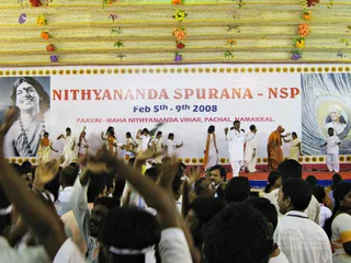 A photograph of a person Spurana convention stage in Namakkal, Tamil Nadu, under a ceiling decorated with garlands and flowers. A large banner reads NITHYANANDA SPURANA - NSP with dates and venue, flanked by black-and-white portraits. On stage, a group of women in white and cream saris with orange borders perform a dance sequence, while a man in white kurta and white shawl with an orange scarf stands center stage with arms raised. Two figures in orange robes stand among them. In the foreground, a dense crowd of audience members, many with raised hands, watches the performance.