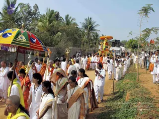 A festival procession moves along a dirt road lined with palm trees and a low wall. At left, a large multicolored canopy with a trident and bird motifs is held aloft on a pole. Behind it, a decorated float or chariot covered in flowers and an orange canopy is pulled forward. Many devotees walk in the procession: men in white kurtas and dhotis, women in white sarees with gold borders, several in orange or saffron robes, and some wearing rudraksha-style beaded necklaces. A few people in the foreground fold their hands in greeting. Vehicles and speakers appear in the background.