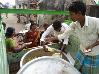 A photograph of a food distribution at a village street in Kuttiyankuppam. A green-painted cart with wooden slats holds two large aluminium vessels filled with cooked food. A young man in a white shirt and dark trousers ladles food onto a plate, while a man in a checked shirt and plaid lungi hands out small paper packets. Women in saris, including one in a red sari with yellow floral print and one in a green blouse, hold out plates to receive food. A bicycle and a few people stand in the background along a dusty road beside weathered houses. No a person candidate is visible.