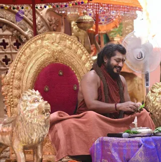 A seated man in rust-orange robes sits on an ornate gold throne with a red velvet back, flanked by gilded lion sculptures. He wears a long beaded necklace, a red sacred thread on his wrist and a vertical tilak on his forehead, and he smiles with eyes closed while holding green leaves in his hands. Beside him on a purple brocade-draped table are a silver vessel, a small silver puja implement and more leaves. A white pedestal fan and an orange and gold canopy with beaded fringe surround the throne. Swamiji (probable): long dark hair, a full beard and a rudraksha-style necklace on a saffron-orange robe, seated on a throne.