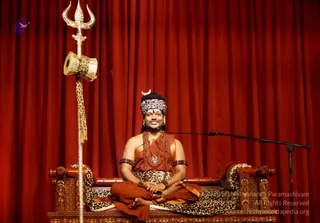 A seated man in orange-red robes sits cross-legged on an ornate wooden throne with leopard-print cushions, in front of a deep red curtain. He wears a leopard-print headband with a silver crescent, rudraksha malas and a silver pendant, with orange bracelets on his wrists. A tall silver trident with a leopard-print drum hangs from it stands beside him, and a microphone stand is at his right. a person: the crescent headband, rudraksha malas and ornate orange clothing match his presentation, and this is an in-person seated satsang scene.