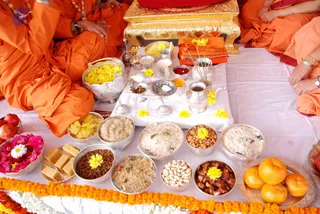 A photograph of a ritual offering spread on a white cloth, viewed from above. Three people in orange robes sit at the edges; only their lap, arms and hands are visible, and one wears gold bangles and a wristwatch. An ornate gold-coloured stand holds a red cushion. Silver bowls and vessels hold rice dishes, sweets, nuts, dried fruit, fresh oranges, apples and bananas, as well as a kumkum dish and a silver pot. Yellow and orange marigold garlands and loose flowers decorate the cloth, with a bowl of pink and white flowers at left. Yellow marigold strands and folded orange cloth lie nearby. The arrangement suggests a naivedyam offering, but no deity form is visible.