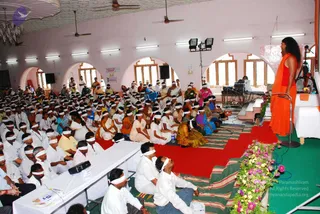 Indoor hall with arched windows and white ceiling fans where a large seated audience of several hundred, many wearing white headbands, sits on striped mats and red carpet. A woman in an orange sleeveless top and dark hair stands at right on a green carpeted stage speaking into a microphone on a stand, beside a white draped table with flower arrangements and sound equipment. Several rows of seated people wear white clothing, some in saffron, pink and blue saris, and a few in white shirts sit at a long white table in front. Stage lights hang overhead and flowers lie at the foot of the stage.
