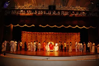 A stage in a concert hall with a dark red curtain drape edged in gold, and a hanging backdrop of orange and yellow fabric decorated with garlands. A seated figure in a red robe sits on an ornate gold throne at centre stage. Around the throne, a long line of people in white clothing and some in saffron and sari attire stand in an arc, several holding items. A person in orange clothing stands near the throne. Overhead lighting rigs and a banner reading Sir Mutha Venkatasubba Rao Concert Hall are visible. The swami figure seated on the throne in red matches the page's description of a person presence, based on his red robe and the throne setting.