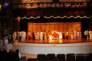 A wide photograph of a stage in a theatre hall during an evening darshan event. A seated figure in red and orange robes sits on an ornate gold-and-red throne at centre stage, in front of a backdrop of hanging orange and gold drapes decorated with marigold-style garlands. A line of about 20 people in white and cream clothes, several women in saris, stands in a semicircle before the throne, some with hands folded or offering items. Dark red curtains and gold valance frame the stage, and an audience sits in rows of seats in the foreground. A small framed portrait hangs at the left side of the stage. The page identifies the event as a Kalpataru darshan, but the seated figure's appearance is not clear enough to identify a candidate.