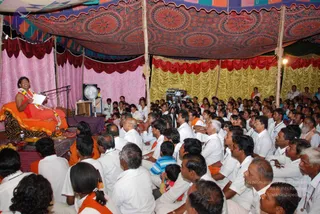 A photograph of a large open-air satsang under a patterned canopy with pink, red and gold draped backdrops. A woman seated on a raised orange-and-black cushioned seat wears an orange sari with a garland and holds a sheet of paper while speaking into a microphone. Before her sit a dense crowd of devotees, mostly men and women in white clothes, several with orange shawls or saffron sashes. A video camera on a tripod records the gathering, and a pedestal fan stands near the speaker. Brass items and offerings sit on a cloth in the foreground. Devotees sit cross-legged on the ground filling the frame.