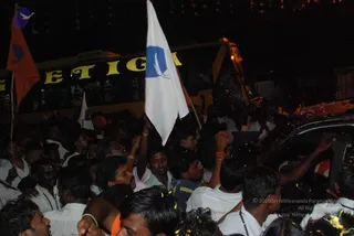 Night photograph of a dense crowd of men gathered in a street near a yellow-lettered bus. Many wear white shirts and white dhotis, some with rudraksha malas and bangles. A large white flag with a blue emblem of a bird is raised high, with an orange flag at left. A dark car at right is surrounded by hands reaching toward it, with scattered yellow flower petals on the vehicle and ground. Strings of decorative flowers hang overhead.