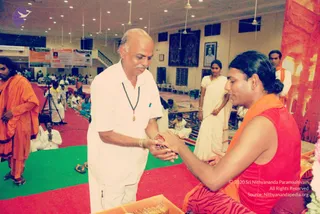 Indoor hall photograph of a ritual exchange on a red carpet with green edging. In the foreground, a man in a white shirt with a dark bead mala and pendant stands and offers items cupped in his hands to a seated man in a sleeveless red top and orange shawl, who receives them over a small cardboard box. To the left, a standing person in orange robes stands beside a tripod. Behind, a woman in white sari stands near a table, and several seated people in white and coloured clothes sit on the floor. Ceiling fans, banners on the upper left wall, framed portraits and a red-orange decorative pillar complete the setting.