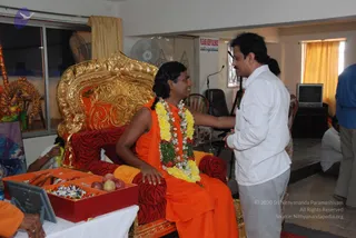 A photograph inside a hall where a long-haired man in saffron-orange robes sits on an ornate gold-and-red throne with carved gilded backrest and a red zebra-striped cushion. He wears a long garland of yellow and white flowers with red blossoms. A man in a white shirt and grey trousers stands beside him, leaning in and touching the seated man's shoulder. On a white table in front, a red tray holds apples, sweets and other offerings, and a framed picture sits beside it. A notice on the wall reads Please Keep Silence, and a tripod microphone and a television stand are visible. Other people in orange and white sit at the edges of the room.