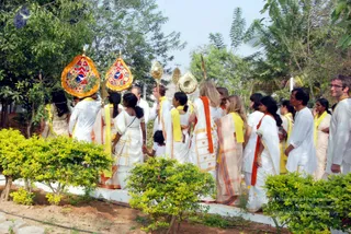 A procession moves along a garden path lined with trimmed shrubs, palms and leafy trees. At front, two attendants carry ornate kalasha-shaped standards with red, orange and gold embroidered panels, and gilt ceremonial staffs topped with gold finials. Participants wear white garments with yellow shawls or sashes, and several women wear orange-bordered white saris. A child in white holds an adult's hand. Among them, a man in white kurta stands at the right edge, wearing a yellow sash. No figure is clearly identifiable as a central presence in this view.
