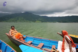 A photograph taken on a green lake under a heavy grey cloud sky, with forested hills behind. In the foreground a blue wooden rowboat holds a smiling woman in sunglasses and an orange life vest, reclining with her bare feet over the side and wearing an orange sari-style wrap. To the right, a second person in an orange headcloth and white dhoti-style clothing sits in the boat, gesturing toward the water. Another blue boat sits in the lower left foreground.