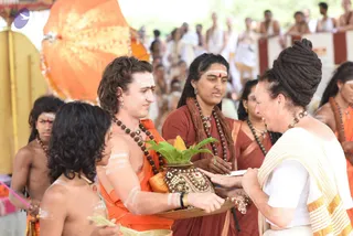 A photograph of an outdoor ceremony with a crowd of onlookers behind. In the centre, a young man with curly brown hair wears a saffron-orange cloth, a rudraksha mala and white sacred markings on his face and arms; he holds a brass tray with a green leaf-decorated kalasha topped with a yellow coconut-like fruit, wrapped in an ornate gold-embroidered cloth. Opposite him, a woman with a large dark topknot wears a white and gold-bordered shawl and rudraksha beads and smiles as she reaches toward the tray. Behind them a woman in a maroon sari with a long beaded mala stands near a shirtless boy in white markings, and an orange-draped canopy forms the backdrop.