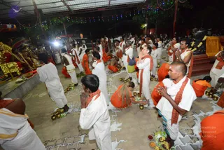 Night photograph of a temple courtyard where many devotees stand and kneel in rows on a paved floor, some with hands folded in namaskar. Many men wear white dhotis and white shirts with orange-red shawls draped over their shoulders, while others wear orange-saffron robes. Brass vessels and small pots are set on the ground, with plates of flowers, fruit and offerings nearby. On the left, a seated figure sits on a gold-decorated throne-like seat with ornate red cushions, flanked by attendants. Strings of coloured lights and a corrugated roof frame the scene, and a black stone lingam draped with a yellow cloth and garlands is visible on the right.