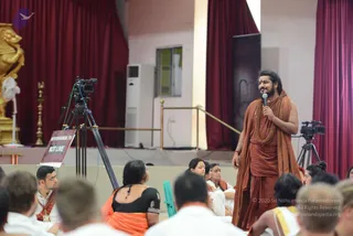 A photograph of an indoor satsang hall at Adi Kailasa, Bengaluru, with red curtains and a golden deity murti at the left edge. A man with long swept-back dark hair and a beard stands on a raised platform holding a microphone, wearing saffron-orange robes and a rudraksha mala, with an orange shawl draped over his shoulder; this appearance matches the Swamiji (probable) cues of long hair, saffron clothing and rudraksha beads, and he is speaking to the seated audience. Seated devotees in front, several in white and saffron garments, face him. A video camera on a tripod is labelled NITHYANANDA TV NOT LIVE, and a second camera sits at right.