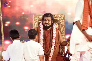 A bearded man with long swept-back dark hair and a red tilak on his forehead sits on an ornate gold-framed throne, wearing a rust-brown robe and a shawl, with a pink and white flower garland and layered rudraksha malas around his neck. Two boys in white shirts sit with their backs to the camera in front of him, and a standing person in white clothing with an orange-striped shawl stands at the right edge. A tall gold staff is visible at the left. Behind them is a glowing red and pink backdrop. Swamiji (probable) is seated on the throne, identified by long swept-back hair, rudraksha malas, ornate gold throne and shawl cues.