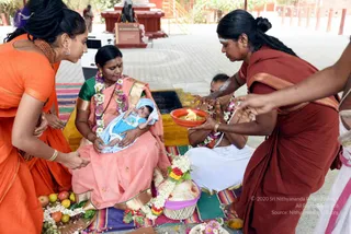 A naming ceremony at an outdoor temple courtyard. A woman in a pink saree with gold border and green blouse, wearing a floral garland, sits holding a swaddled infant in blue. Beside her, a woman in an orange saree and orange-beaded jewelry leans in. On the right, a woman in a maroon saree with gold border and white tilak on her forehead holds a small orange bowl with a flame over the baby's area, while another woman in white sits behind, garlanded. Fruits, flowers, a woven basket and a banana leaf with offerings lie on colourful striped mats. Red-painted structures and a plaque stand in the background.