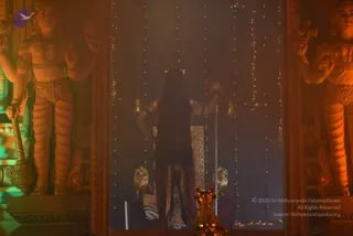 A photograph of a dimly lit shrine interior with warm orange light and haze. A figure seen from behind stands centrally with long dark hair falling down the back, wearing a dark brown draped garment and a small crown-like headpiece, with arms raised and hands gripping hanging chains or rods. Strings of small lights and hanging brass bells frame the figure. Two tall sculpted guardian figures with multiple arms flank the frame on either side, and a golden lamp stand with a lit flame sits in the lower foreground. A tiger-skin cloth is visible beneath the figure, and a golden ornate seat or throne stands behind.