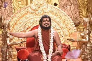 A photo of a seated man on an ornate golden throne with a red cushioned seat, carved gilded figures flanking him on both sides. He wears a rust-red sannyasi robe, a long white and pink flower garland, a gold chain, and a red tilak on his forehead; his left arm is extended outward and he sits barefoot with a smile. Swamiji (probable): the long swept-back hair, ornate gold throne setting, red-saffron robe and flower garland are consistent with the page's reference to Swamiji. One person is visible.