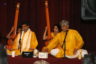 Four performers sit cross-legged on a red-draped stage in front of a dark red curtain, playing devotional music before a seated audience whose heads are visible at the bottom edge. Two men at the front wear saffron-yellow silk kurtas and white dhotis, each with a microphone; the man on the left has a cream shawl with an orange border, and the man on the right plays a small Raagini Digital harmonium-style box. Behind them, two women in saffron-yellow clothing hold long wooden tanpura-type stringed instruments, and a young girl with glasses sits at the left edge beside another instrument. Paper cups sit on the stage, and a Radel speaker is on the floor.
