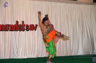 A performer on a stage dances in a dramatic pose, one arm raised high and one leg lifted behind him. He wears a black crown-like headpiece with a red cloth draped behind, a bare chest with metal and beaded necklaces, a leopard-print waistcloth over orange trousers, a green dhoti-style skirt with gold trim, and gold and beaded anklets and armbands. A white draped backdrop behind him bears red lettering reading 'mandotsav', and a green stage floor is visible below. The performer is a dancer in costume, with no clear identification of a specific person.