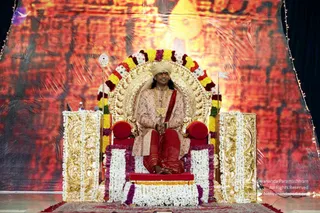 A seated man on an ornate gold throne (peetha) framed by a garland arch of yellow, white, red and purple flowers. He wears a cream turban, a cream and pink embroidered long kurta with gold chains, and red dhoti-style trousers, with hands folded in his lap. White and purple flower garlands drape the gold side pillars and the red-cushioned platform. Behind him is a large backdrop of warm orange-red tones with a glowing golden figure, and string lights line the edges. A small water bottle and microphone sit on the left. The man's visible cues, including the turban, ornate clothing and throne setting, make him a probable Swamiji seated in the role of a presiding teacher.