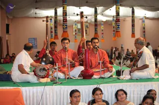 A musical performance on a green-carpeted stage under white draped ceiling fabric with colourful woven bands hanging from above. Four musicians sit on the stage: a bald man in white on the left plays a small drum with a mallet, two younger men in saffron-orange kurtas and white dhotis sing into microphones, one holding a tanpura-style instrument, and an older man in white on the right plays a violin-like string instrument. A red-painted pot-shaped resonator stands in front. Seated women in saris watch in the foreground and the audience sits on the floor behind. No a person candidate is visible on stage.