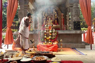 A photograph of an outdoor shrine during a puja. A priest in white dhoti and shawl stands with his hands together before a stone shrine decorated with marigold and orange garlands, gold lamps and brass vessels. Smoke rises from incense in front of the central deity murti and goddess figures. An orange-robed person sits on the left near a low platform. Orange curtains frame the shrine. Trays of offerings in front include fruit, sweets, rice and red flowers, with a small microphone stand nearby. No one is confidently identifiable as a person in this image.