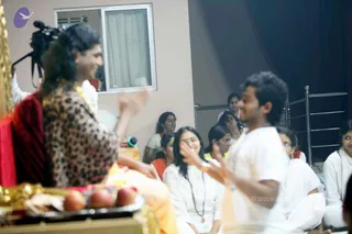 A satsang gathering indoors with a seated figure at left wearing a red garment, a leopard-print patterned top and gold necklaces, gesturing with a raised hand; a plate of red apples sits in the foreground. A man in a white shirt at right gestures animatedly. Behind them, several women in white and orange-red clothing sit on the floor, one wearing a rudraksha-style mala, some smiling. A video camera on a tripod stands at upper left, and a window with a curtain and a metal rack are visible in the room.